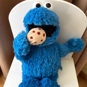 Könnte beinhalten: Ein blaues Cookie Monster-Plüschtier mit großen schwarzen Augen und offenem Mund, das einen Schokoladenkeks hält. Das Spielzeug sitzt auf einem weißen Stuhl.