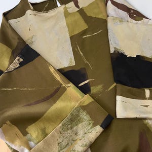 May include: A long-sleeved blouse with a round neckline and a keyhole back. The fabric features an abstract pattern in shades of olive green, beige, black, and brown. The blouse has a relaxed fit and is made of a smooth, flowing material.