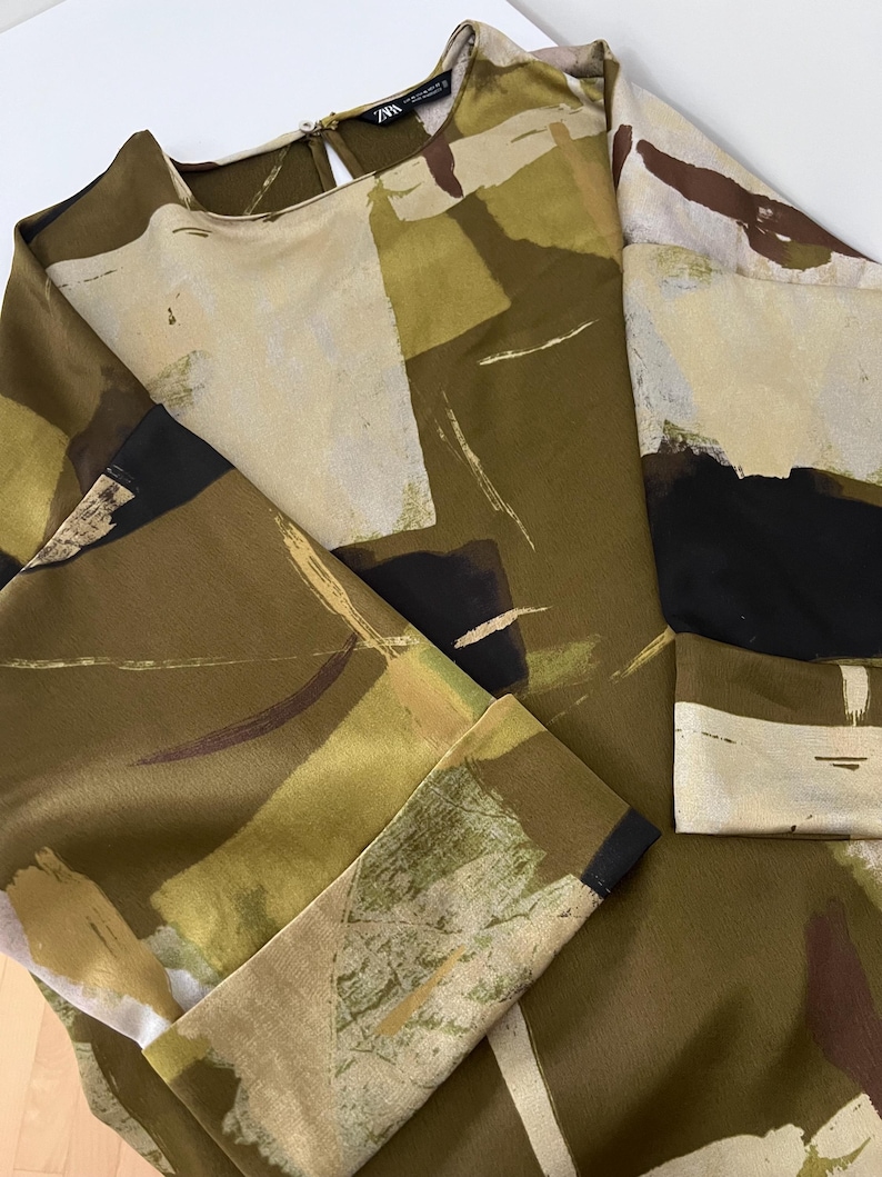 May include: A long-sleeved blouse with a round neckline and a keyhole back. The fabric features an abstract pattern in shades of olive green, beige, black, and brown. The blouse has a relaxed fit and is made of a smooth, flowing material.