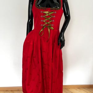 May include: A red dirndl dress with a square neckline and a fitted bodice. The dress features a green ribbon corset, brown trim, and a full skirt. The dress is displayed on a black mannequin.