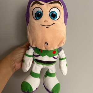 Pode incluir: Um boneco de pel&uacute;cia Buzz Lightyear, com cabe&ccedil;a roxa, rosto rosa claro e olhos azuis. O boneco usa um traje espacial branco e verde com um bot&atilde;o vermelho. Um brinquedo divertido para crian&ccedil;as.