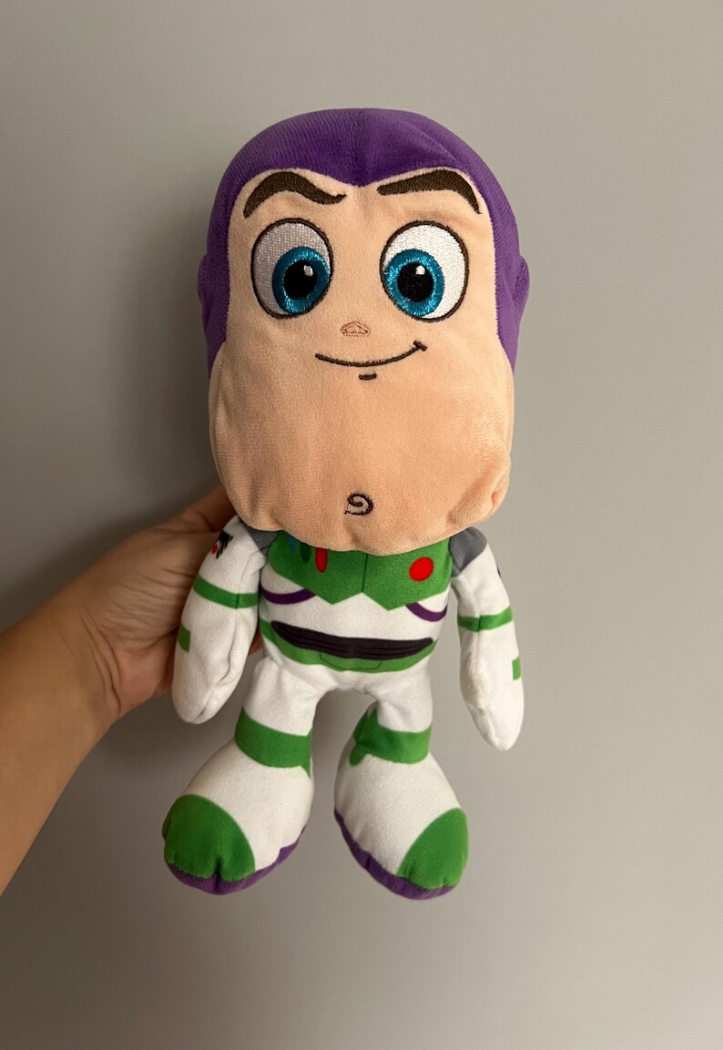 Pode incluir: Um boneco de pel&uacute;cia Buzz Lightyear, com cabe&ccedil;a roxa, rosto rosa claro e olhos azuis. O boneco usa um traje espacial branco e verde com um bot&atilde;o vermelho. Um brinquedo divertido para crian&ccedil;as.
