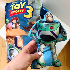 May include: A rectangular pillow featuring the Toy Story 3 logo and characters including Buzz Lightyear, Jessie, and Rex, alongside a separate, smaller, die-cut plush toy shaped like Buzz Lightyear in his signature white, green, and purple suit.