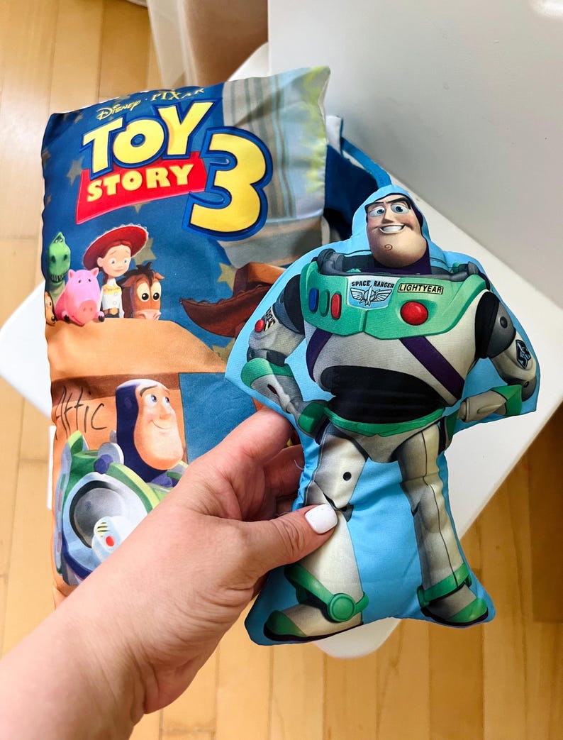 May include: A rectangular pillow featuring the Toy Story 3 logo and characters including Buzz Lightyear, Jessie, and Rex, alongside a separate, smaller, die-cut plush toy shaped like Buzz Lightyear in his signature white, green, and purple suit.