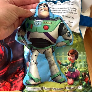 May include: A plush fabric toy shaped like Buzz Lightyear from Toy Story, featuring a printed space ranger suit in white, green, and purple. The toy is layered over a soft book page with printed text and an illustration of a child and toys.