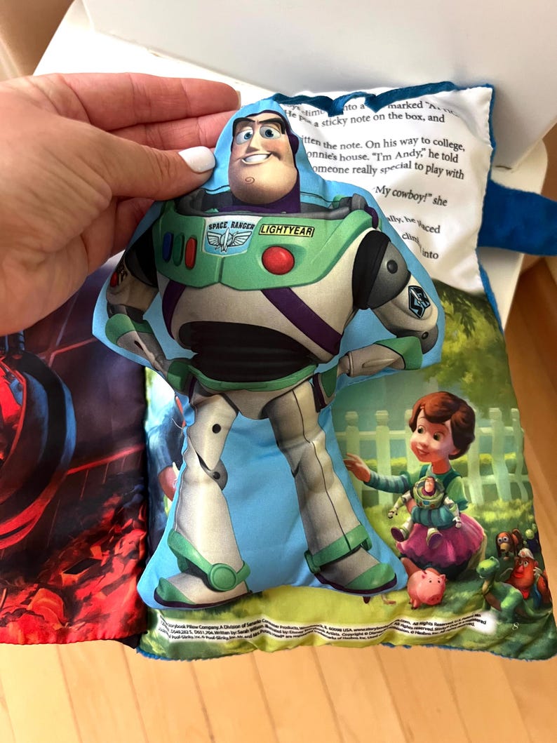 May include: A plush fabric toy shaped like Buzz Lightyear from Toy Story, featuring a printed space ranger suit in white, green, and purple. The toy is layered over a soft book page with printed text and an illustration of a child and toys.