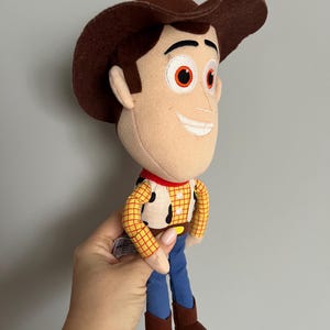 Puede incluir: Un peluche del personaje Woody de la pel&iacute;cula Toy Story. La mu&ntilde;eca tiene un sombrero de vaquero marr&oacute;n, una cara bronceada y una camisa a cuadros amarilla con un pa&ntilde;uelo rojo. Pantalones azules y botas marrones completan el atuendo.