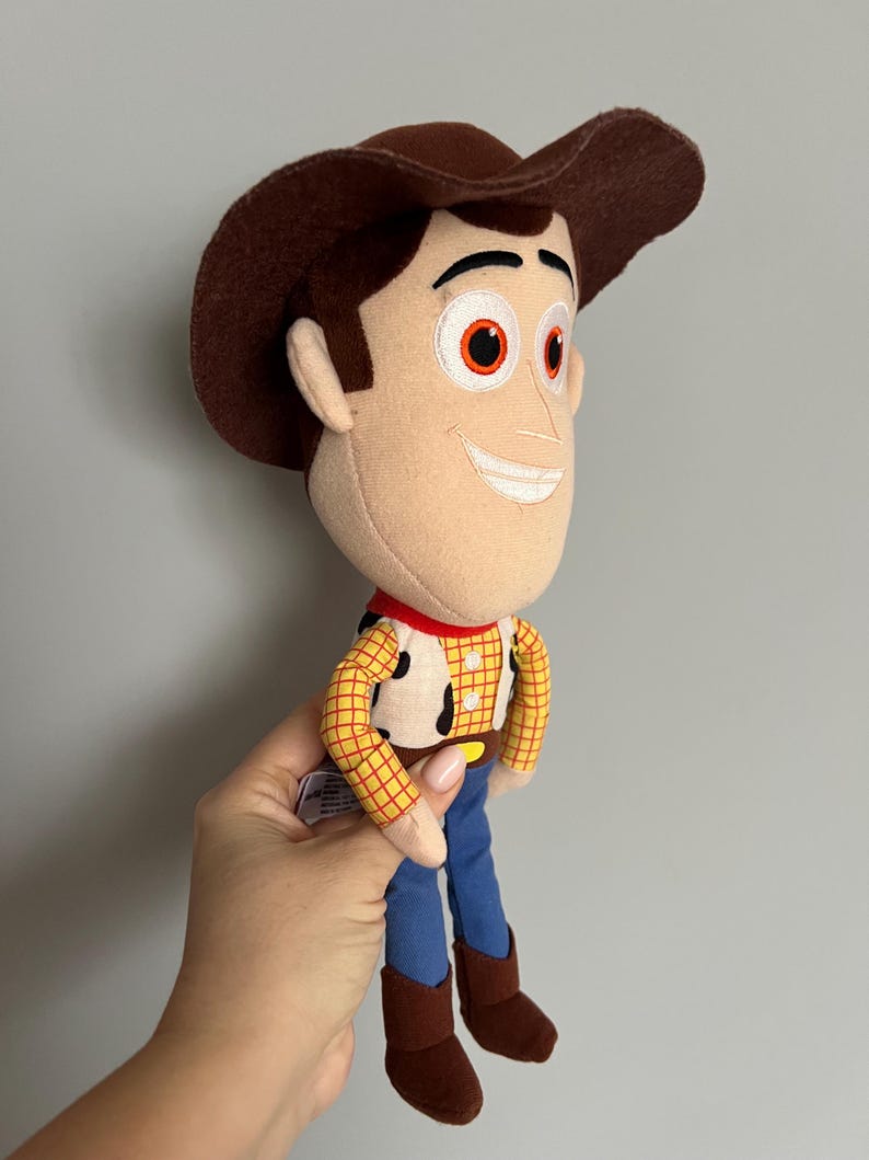 Puede incluir: Un peluche del personaje Woody de la pel&iacute;cula Toy Story. La mu&ntilde;eca tiene un sombrero de vaquero marr&oacute;n, una cara bronceada y una camisa a cuadros amarilla con un pa&ntilde;uelo rojo. Pantalones azules y botas marrones completan el atuendo.