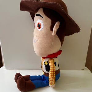 Puede incluir: Un peluche de un personaje de dibujos animados de vaquero, con sombrero marr&oacute;n, pa&ntilde;uelo rojo, camisa a cuadros amarilla, pantalones azules y botas marrones. El juguete est&aacute; sentado.