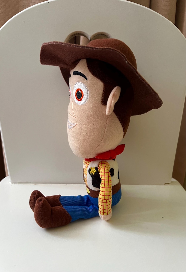 Puede incluir: Un peluche de un personaje de dibujos animados de vaquero, con sombrero marr&oacute;n, pa&ntilde;uelo rojo, camisa a cuadros amarilla, pantalones azules y botas marrones. El juguete est&aacute; sentado.