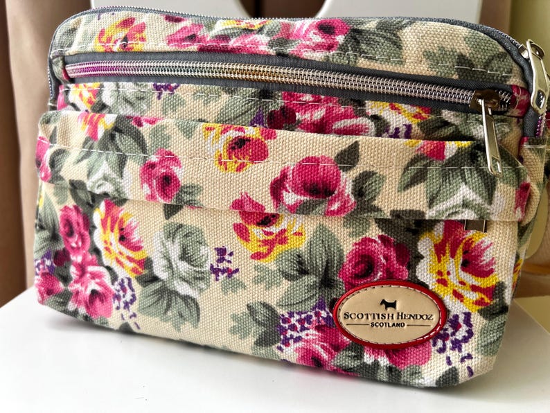 Op de afbeelding: Crossbody tas met een bloemenpatroon met roze, gele en witte rozen op een beige achtergrond. De tas heeft een grijze rits en een klein voorvak. Een leren patch met de tekst "Scottish Henndz Scotland" staat op de voorkant.