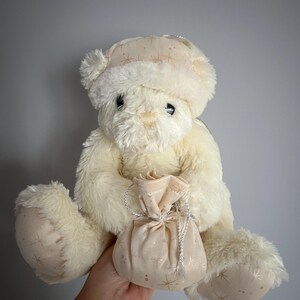 May include: Cream-colored teddy bear wearing a matching hat with gold star patterns. The bear is holding a small, cream-colored pouch with a silver drawstring. The plush toy has soft fur and is designed for children.