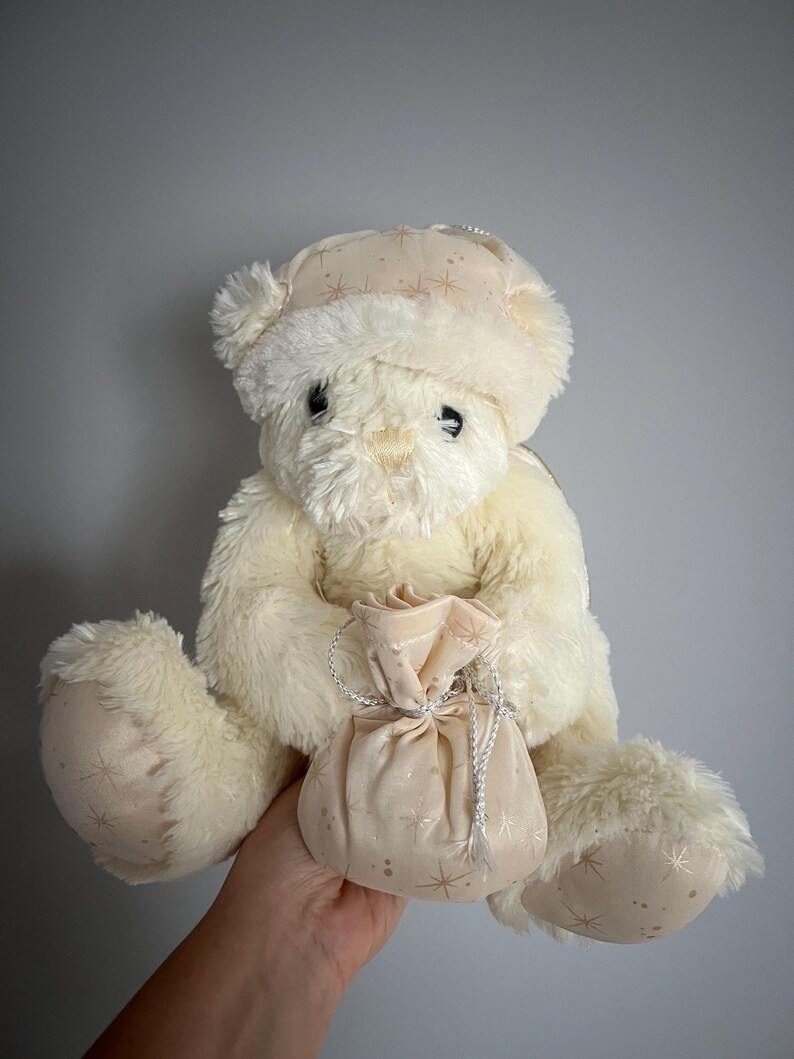 May include: Cream-colored teddy bear wearing a matching hat with gold star patterns. The bear is holding a small, cream-colored pouch with a silver drawstring. The plush toy has soft fur and is designed for children.