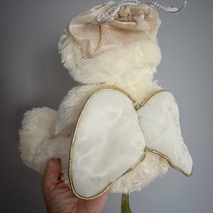 May include: A plush, cream-colored angel doll with a soft, fluffy body and a matching hat. The doll features large, white wings with gold trim and a delicate star pattern. A silver drawstring bag is attached to the hat.