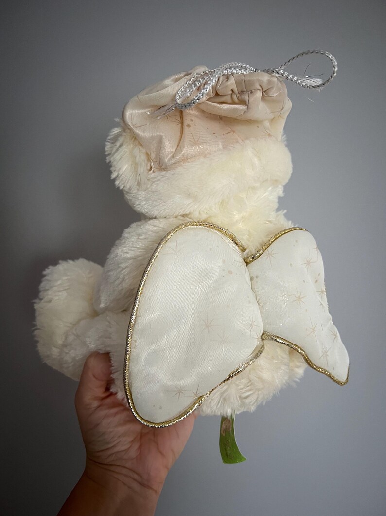 May include: A plush, cream-colored angel doll with a soft, fluffy body and a matching hat. The doll features large, white wings with gold trim and a delicate star pattern. A silver drawstring bag is attached to the hat.