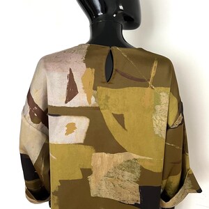 May include: A long-sleeved blouse with a unique abstract pattern. The blouse features a keyhole detail at the back and is adorned with a mix of olive green, beige, brown, and white geometric shapes. The sleeves are cuffed at the wrists.