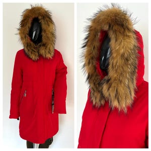 Puede incluir: Un abrigo de invierno rojo vibrante con una gran capucha adornada con piel marrón y negra. El abrigo tiene cierre de cremallera y un diseño elegante y moderno. El abrigo se exhibe en un maniquí.