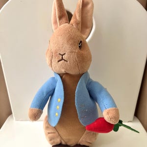 Puede incluir: Un peluche de conejo con pelaje marrón, que lleva una chaqueta azul claro con tres botones amarillos. El conejo sostiene una zanahoria roja con hojas verdes. El juguete está sentado en una silla blanca.