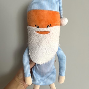 Op de afbeelding: Een pluche wortelspeelgoed met een blauw-wit gestreepte nachtmuts en pyjama. De wortel heeft een witte baard en een lachend gezicht. Het speelgoed is zacht en knuffelbaar.