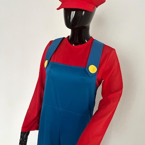 Puede incluir: Un sombrero rojo con un logotipo "M" blanco, una camisa roja de manga larga y un mono azul con botones amarillos. El disfraz se exhibe en un maniquí negro. Este atuendo está inspirado en un popular personaje de videojuego.