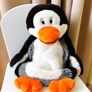 Op de afbeelding: Een pluche pinguïn met een zwarte kop en vleugels, een witte snuit en buik en oranje voeten en snavel. De pinguïn zit op een witte stoel. Het speelgoed heeft een zachte, donzige textuur.