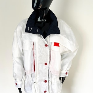 Op de afbeelding: Witte jas met een marineblauwe kraag en rode accenten. De jas heeft rode knopen, zakdetails en afwerking. De jas wordt getoond op een zwarte mannequin.