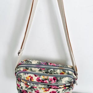 Op de afbeelding: Crossbody tas met bloemenprint met een beige achtergrond en een cr&egrave;mekleurige riem. De tas heeft een patroon van roze en gele rozen met groene bladeren. Het heeft drie compartimenten met ritssluiting.