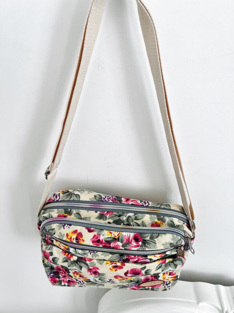 Op de afbeelding: Crossbody tas met bloemenprint met een beige achtergrond en een cr&egrave;mekleurige riem. De tas heeft een patroon van roze en gele rozen met groene bladeren. Het heeft drie compartimenten met ritssluiting.