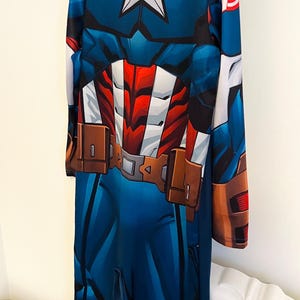 May include: Blue Captain America costume with a white star on the chest, red and white accents, and brown belt details. The long-sleeved jumpsuit features a zipper closure and is designed to resemble the superhero's uniform.