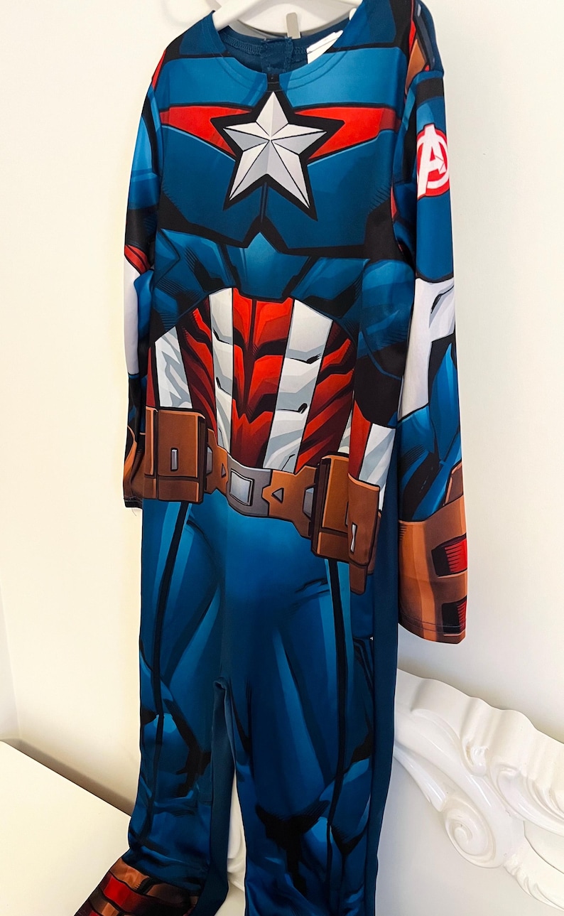 May include: Blue Captain America costume with a white star on the chest, red and white accents, and brown belt details. The long-sleeved jumpsuit features a zipper closure and is designed to resemble the superhero's uniform.