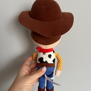 Puede incluir: Un peluche de un personaje de vaquero, con sombrero marr&oacute;n, pa&ntilde;uelo rojo, chaleco con estampado de vaca, pantalones azules y botas marrones. El juguete tiene mangas de cuadros amarillos y una etiqueta que dice "Disney Pixar".
