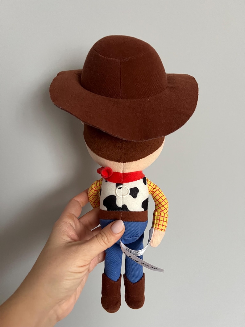 Puede incluir: Un peluche de un personaje de vaquero, con sombrero marr&oacute;n, pa&ntilde;uelo rojo, chaleco con estampado de vaca, pantalones azules y botas marrones. El juguete tiene mangas de cuadros amarillos y una etiqueta que dice "Disney Pixar".