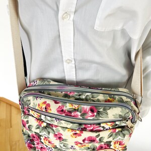 Op de afbeelding: Crossbody tas met bloemenprint en meerdere compartimenten met ritssluiting. De tas heeft een cr&egrave;mekleurige achtergrond met roze en rode rozen, groene bladeren en een verstelbare beige riem. De ritsen zijn grijs en de tas heeft een klein bruin label.
