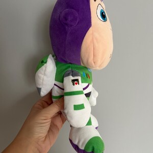 Pode incluir: Boneco de pel&uacute;cia Buzz Lightyear, com cabe&ccedil;a e capacete roxos, rosto cor de p&ecirc;ssego e roupa espacial verde e branca. O boneco tem tra&ccedil;os faciais detalhados e &eacute; feito de tecido macio e texturizado. O brinquedo &eacute; um personagem da franquia Toy Story.