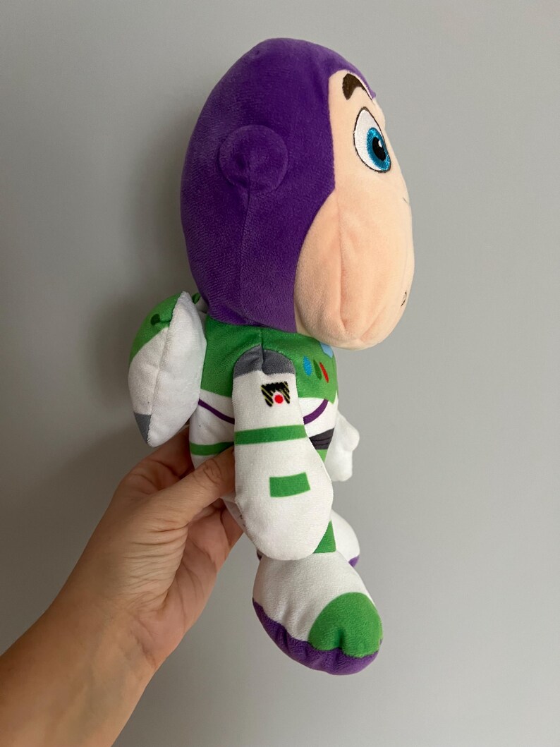 Pode incluir: Boneco de pel&uacute;cia Buzz Lightyear, com cabe&ccedil;a e capacete roxos, rosto cor de p&ecirc;ssego e roupa espacial verde e branca. O boneco tem tra&ccedil;os faciais detalhados e &eacute; feito de tecido macio e texturizado. O brinquedo &eacute; um personagem da franquia Toy Story.
