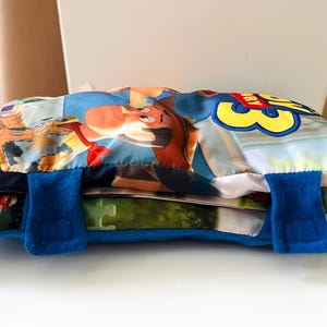 May include: A rectangular Toy Story 3 themed fabric pouch featuring a colorful graphic of Woody on the front. The pouch is secured with two blue felt straps with hook-and-loop fasteners. The design includes the Toy Story 3 logo in yellow and red.