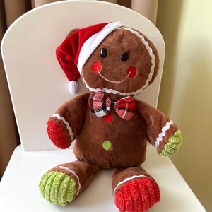 May include: A soft gingerbread man toy wearing a red and white Santa hat and a plaid bow tie. The gingerbread man has a smiling face with black button eyes and red cheeks. Its feet are red and green, and it is sitting on a white chair.