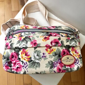 Op de afbeelding: Crossbody tas met bloemenprint op een beige achtergrond, met roze, gele en paarse bloemen. De tas heeft een beige band en een ritssluiting. Een patch met de tekst "Scottish Hendoz - Scotland" is zichtbaar.