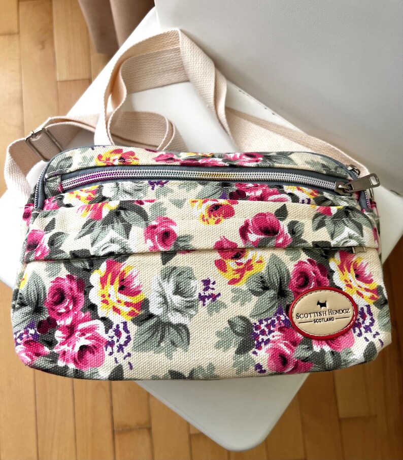 Op de afbeelding: Crossbody tas met bloemenprint op een beige achtergrond, met roze, gele en paarse bloemen. De tas heeft een beige band en een ritssluiting. Een patch met de tekst "Scottish Hendoz - Scotland" is zichtbaar.