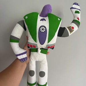 May include: A plush Buzz Lightyear doll, featuring a white body with green, purple, and gray accents. The doll has a raised arm, showcasing details like a red circle and a yellow patch. The design is inspired by the character from the Toy Story movies.