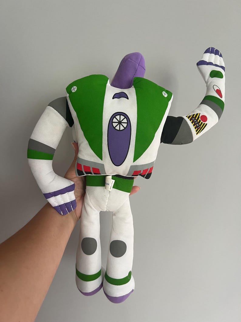 May include: A plush Buzz Lightyear doll, featuring a white body with green, purple, and gray accents. The doll has a raised arm, showcasing details like a red circle and a yellow patch. The design is inspired by the character from the Toy Story movies.