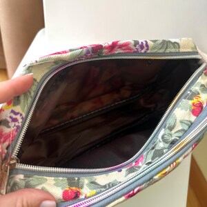Op de afbeelding: Make-uptas met bloemenprint en ritssluiting. De tas heeft een cr&egrave;mekleurige achtergrond met een patroon van roze, paarse en groene bloemen. De binnenkant is donkerbruin. De rits is zilverkleurig en de rand grijs.