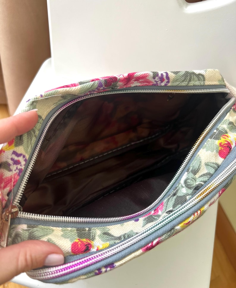 Op de afbeelding: Make-uptas met bloemenprint en ritssluiting. De tas heeft een cr&egrave;mekleurige achtergrond met een patroon van roze, paarse en groene bloemen. De binnenkant is donkerbruin. De rits is zilverkleurig en de rand grijs.