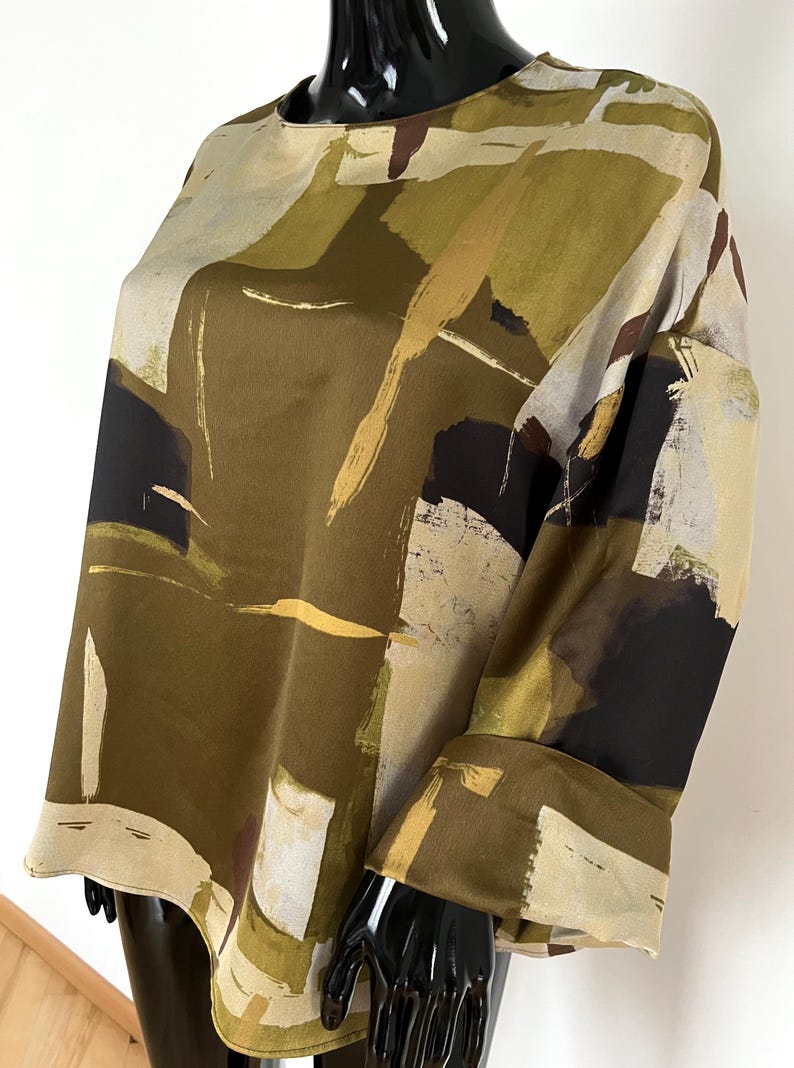 May include: An olive green, beige, and black abstract print blouse with long sleeves. The top has a round neckline and a loose fit. The sleeves have a cuff at the wrist. The fabric has a silky appearance.