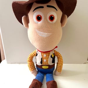Puede incluir: Un peluche de un personaje de vaquero, con sombrero marr&oacute;n, camisa a cuadros amarilla, pantalones azules y botas marrones. El personaje tiene una cara sonriente con detalles bordados y un pa&ntilde;uelo rojo. Una placa de sheriff est&aacute; en el chaleco.