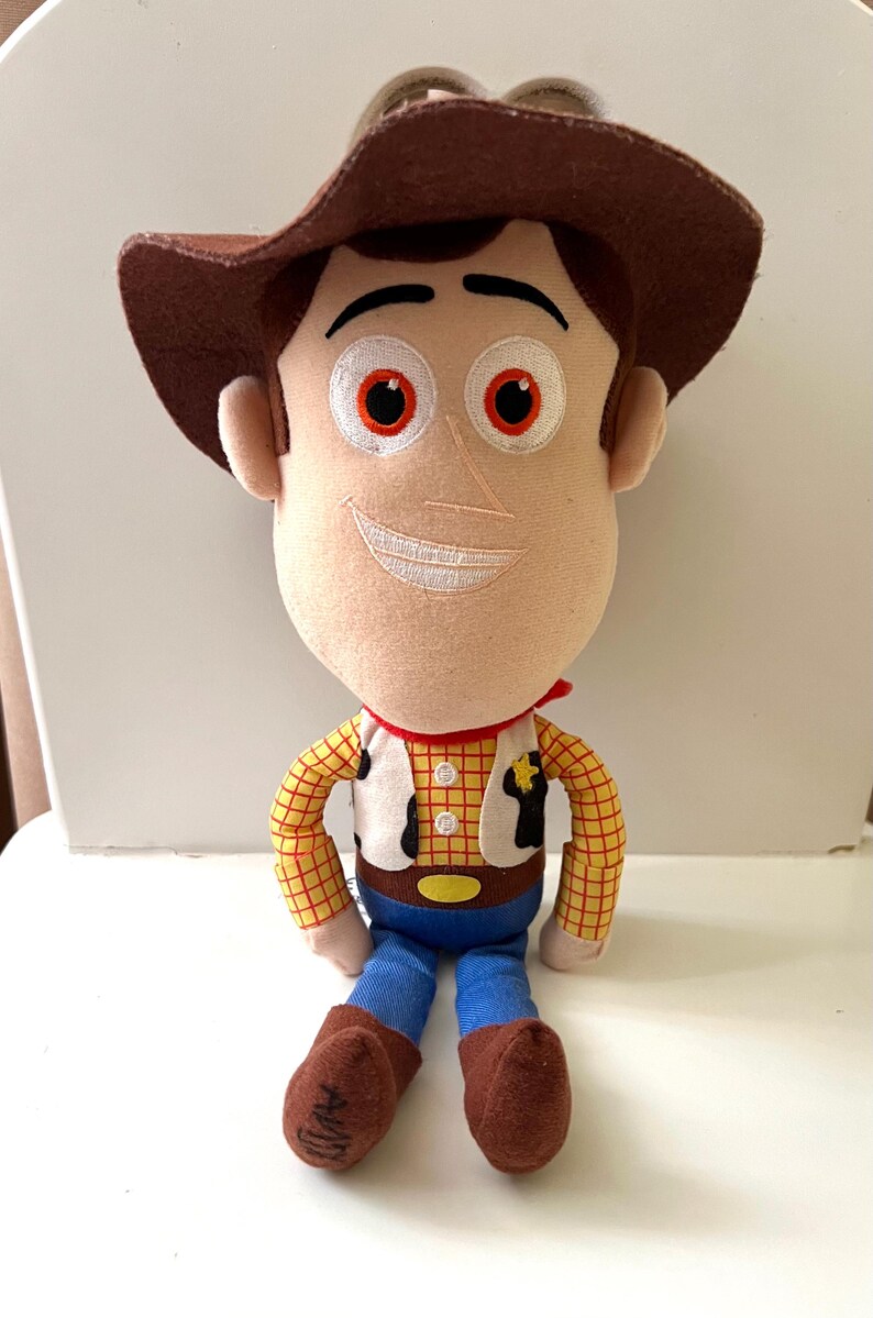 Puede incluir: Un peluche de un personaje de vaquero, con sombrero marr&oacute;n, camisa a cuadros amarilla, pantalones azules y botas marrones. El personaje tiene una cara sonriente con detalles bordados y un pa&ntilde;uelo rojo. Una placa de sheriff est&aacute; en el chaleco.