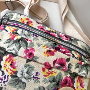 Op de afbeelding: Een crossbodytas met bloemenpatroon met een cr&egrave;mekleurige achtergrond en een patroon van roze, gele en paarse bloemen. De tas heeft een cr&egrave;mekleurige riem en een ritssluiting. Het label luidt "Scottish Hemdoz Scotland."