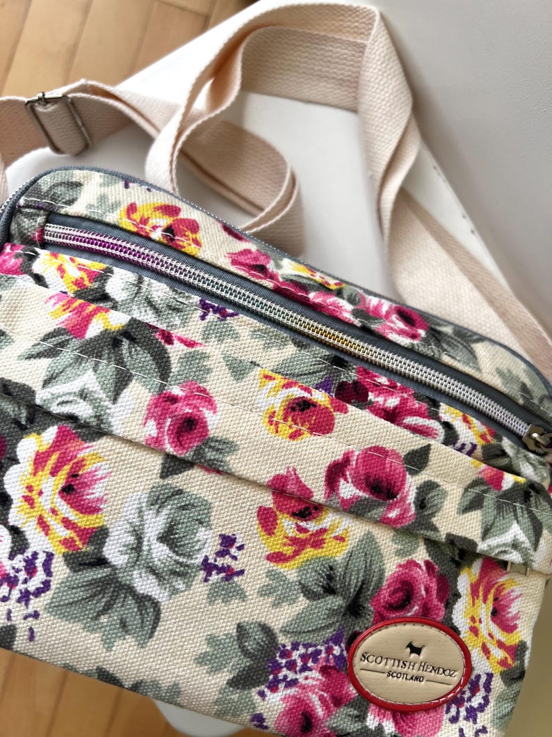 Op de afbeelding: Een crossbodytas met bloemenpatroon met een cr&egrave;mekleurige achtergrond en een patroon van roze, gele en paarse bloemen. De tas heeft een cr&egrave;mekleurige riem en een ritssluiting. Het label luidt "Scottish Hemdoz Scotland."