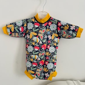Könnte beinhalten: Ein marineblauer Baby-Badeanzug mit einem lebendigen Blumenmuster. Das Muster umfasst rote, gelbe und hellblaue Blumen und Schmetterlinge. Er hat kurze Ärmel und einen hohen Kragen mit gelben Verzierungen.