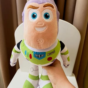 May include: A plush Buzz Lightyear doll, featuring a light purple head and a green and white spacesuit. The doll has large blue eyes, a smiling expression, and the word "LIGHTYEAR" on the chest. The doll is held in front of a white chair.